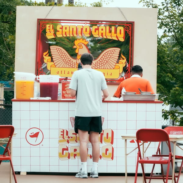 Two men stand at the El Santo Gallo Taqueria food stall with drinks and red chairs.