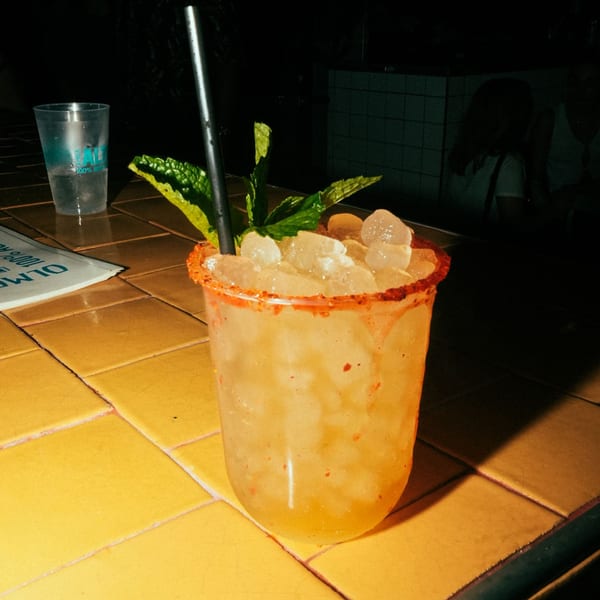 A Mexican cocktail with ice, mint, and a chili-rimmed glass on a yellow tiled surface.