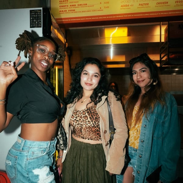 Three young women posing happily in front of a menu board at El Santo Gallo Taqueria.