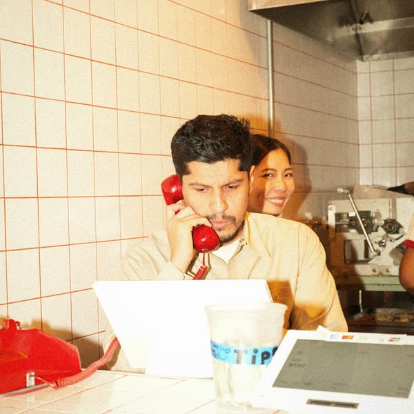 A man on a red phone at a counter, with a smiling woman behind him in a kitchen.