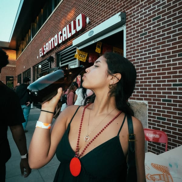 A woman drinks from a large bottle outside El Santo Gallo Taqueria, with brick walls and festive decor.