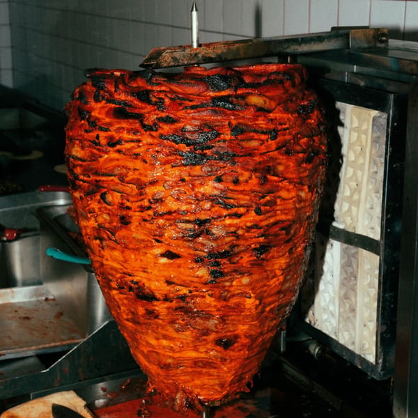 Large vertical rotisserie of marinated al pastor pork cooking, with charred edges, at El Santo Gallo Taqueria.