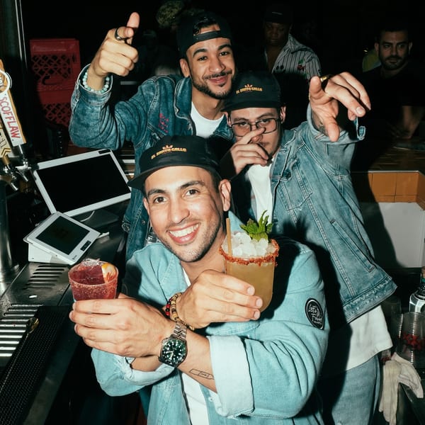 Three men in El Santo Gallo hats and denim jackets holding cocktails at a bar.