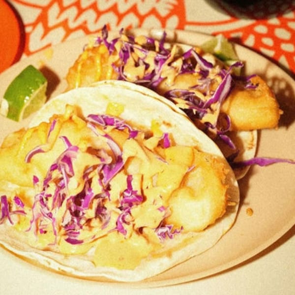 Two El Santo Gallo fish tacos with purple cabbage, sauce, and lime wedges on a plate.