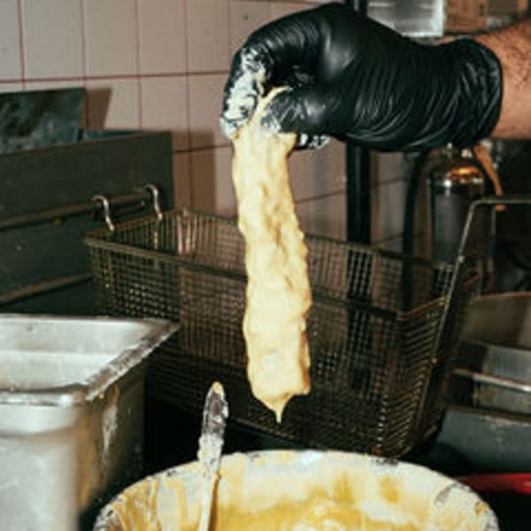 A gloved hand holds a churro covered in batter over a deep fryer basket.