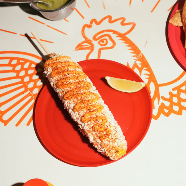 Elote, Mexican street corn, on a red plate with a lime wedge, on a rooster-patterned table.