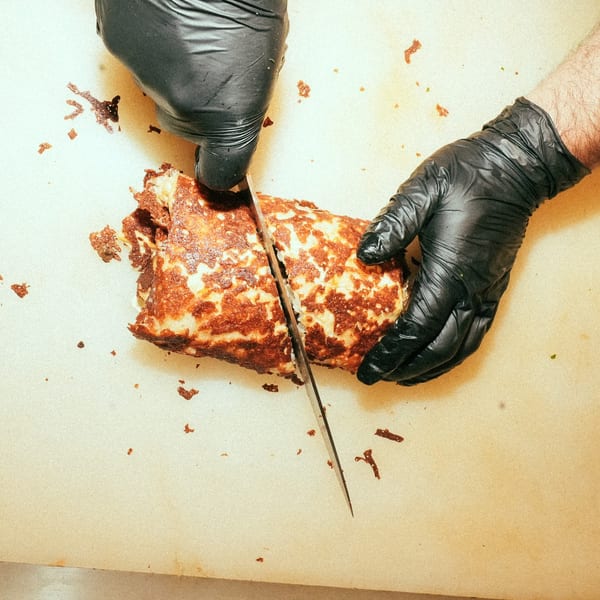 Gloved hands slicing a large, browned, cylindrical Mexican dish on a light surface.