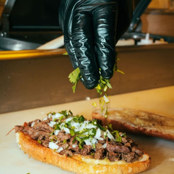 A chef's gloved hand sprinkles cilantro and onions onto a Mexican torta with meat.