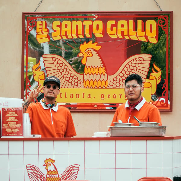 Two men in orange shirts behind a counter with a large El Santo Gallo sign above them.