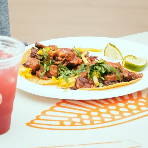 El Santo Gallo Taqueria's carne asada taco with cilantro, pineapple, and lime wedges, next to a red drink.