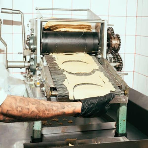 A chef's tattooed arm and gloved hand operating a tortilla press, making fresh El Santo Gallo tortillas.