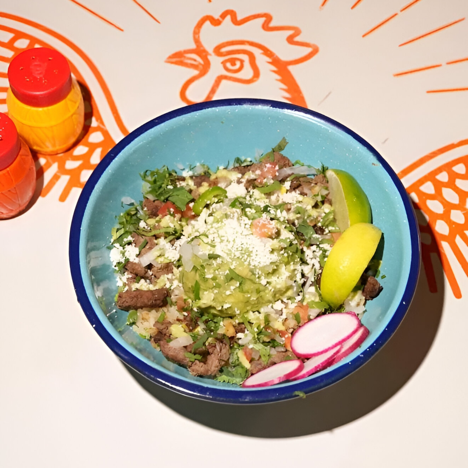 A blue bowl filled with carne asada, guacamole, cheese, and radishes, with lime wedges.