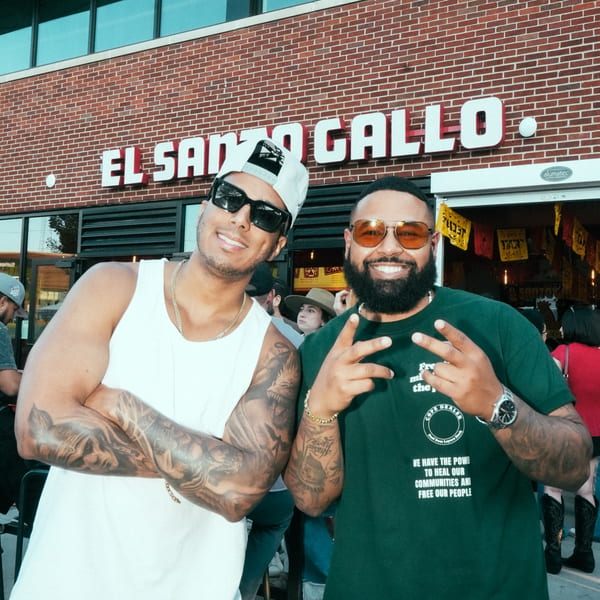 Two men pose smiling in front of El Santo Gallo Taqueria, one making a peace sign.