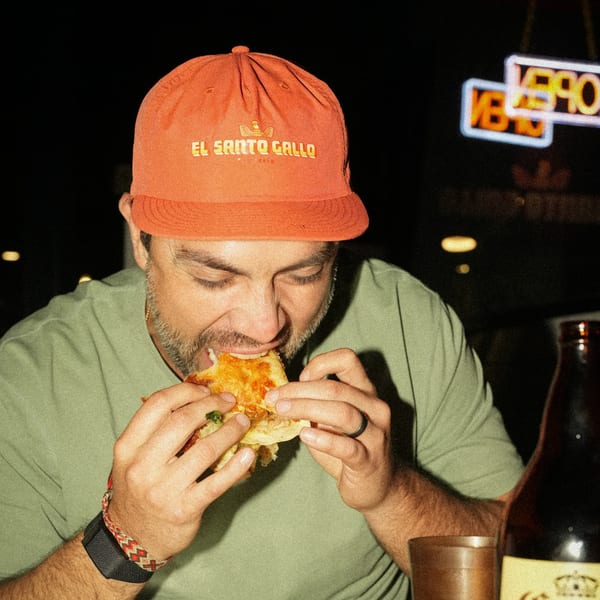 Man in an El Santo Gallo hat enjoying a quesadilla at night, with a neon sign in the background.
