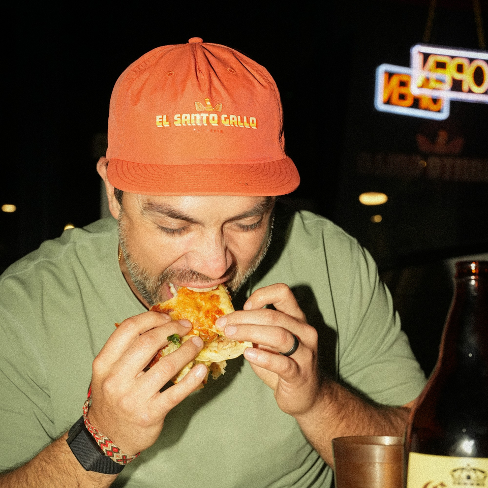 Man in an El Santo Gallo hat enjoying a quesadilla at night, with a neon sign in the background.