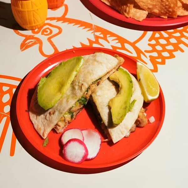 Two El Santo Gallo Taqueria quesadillas with avocado slices, lime, and radishes on a red plate.